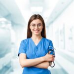 Young nurse in blue scrubs smiling and holding a stethoscope in a hospital corridor.